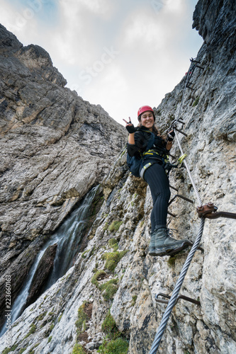 young attractive female mountain climber on a difficult Via Ferrata in the Dolomites in Alta Badia in the South Tyrol in Italy making a peace sign and smiling