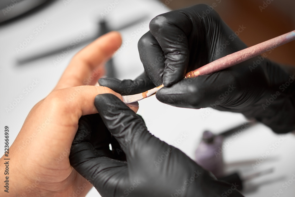 Process of making manicure in salon. Close-up of manicure master hand in black rubber gloves applying with brush transparent gel polish on woman fingernails on blurred background. Fashion and beauty.