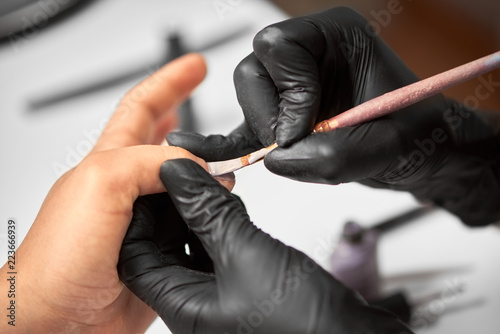 Process of making manicure in salon. Close-up of manicure master hand in black rubber gloves applying with brush transparent gel polish on woman fingernails on blurred background. Fashion and beauty.