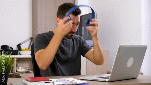 Man types fast on computer keyboard then puts headphones on his head. There is a TV playing a video in the background. Dolly slider 4K footage