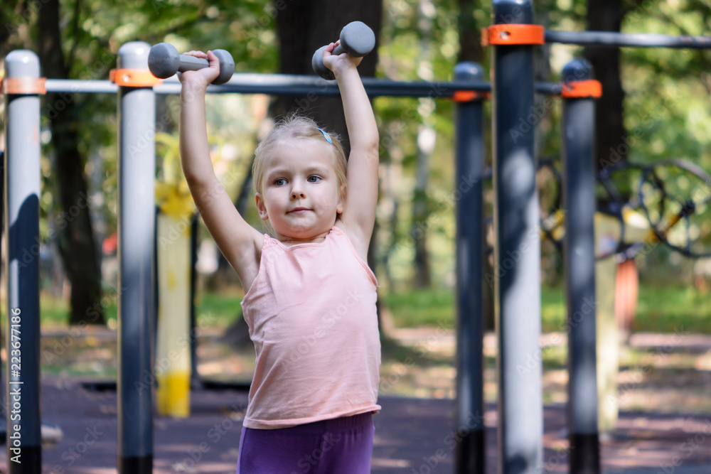 The child high raises dumbbells. A little girl with blond hair goes in for sports. The child high raises dumbbells. A little girl with blond hair goes in for sports.
