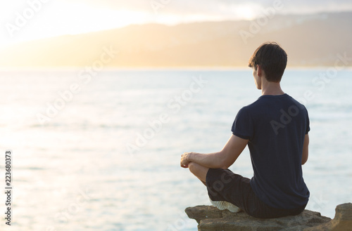 Young man meditating on top ocean cliff during sunset. 