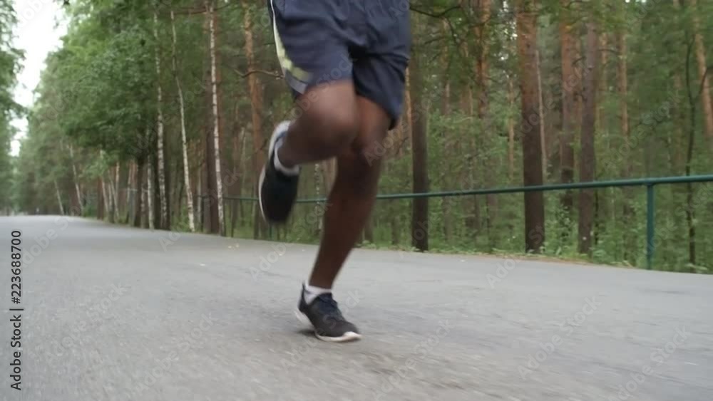 Low-section dolly shot of unrecognizable black man in shorts jogging on pavement in park Low-section dolly shot of unrecognizable black man in shorts jogging on pavement in park
