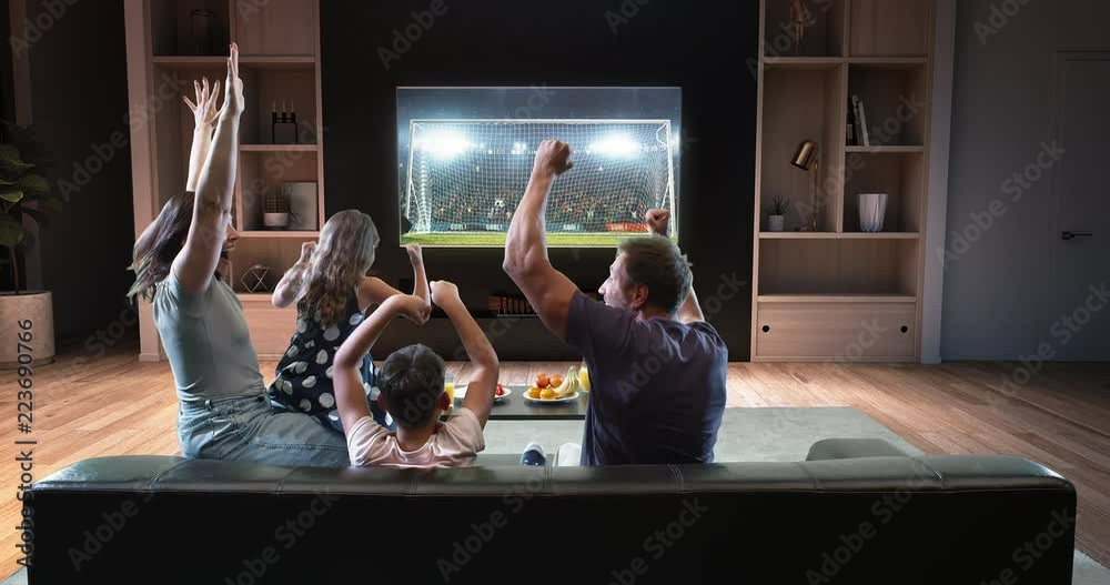 A family is watching a soccer moment on the TV and celebrating a goal, sitting on the couch in the living room. The living room is made in 3D.