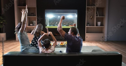 A family is watching a soccer moment on the TV and celebrating a goal, sitting on the couch in the living room. The living room is made in 3D.