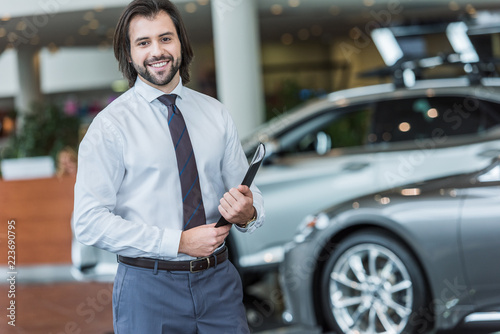 smiling seller with folder standing in dealership salon with cars on background