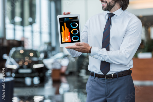 partial view of dealership salon seller in formal wear showing tablet with infographic in hands photo