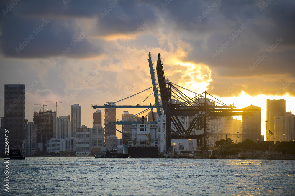 Container ship being unloaded with cranes at a port with city skyline backdrop at sunset