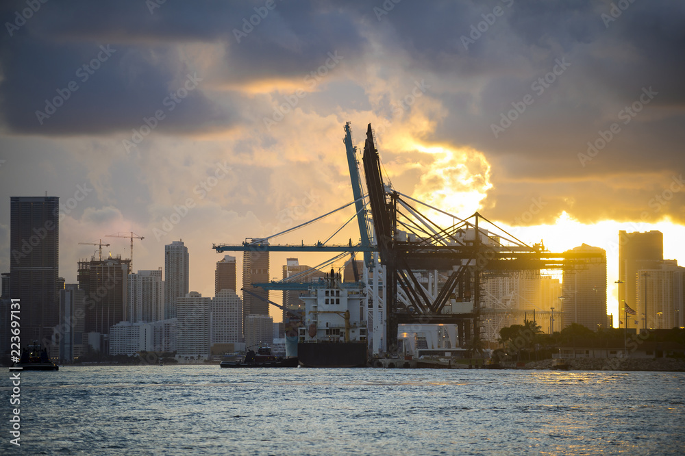 Container ship being unloaded with cranes at a port with city skyline backdrop at sunset Container ship being unloaded with cranes at a port with city skyline backdrop at sunset