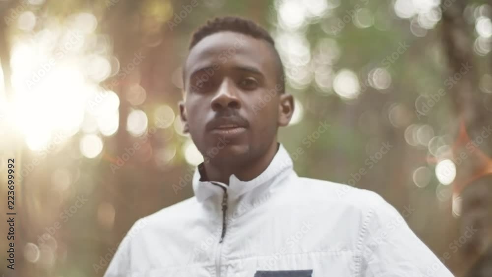Tracking shot of confident black man in windbreaker standing in forest on sunny day and smiling for camera Tracking shot of confident black man in windbreaker standing in forest on sunny day and smiling for camera