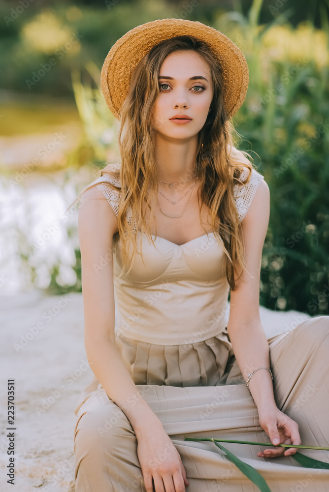 beautiful girl in straw hat sitting on ground near lake