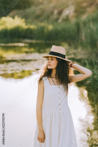 elegant curly girl posing in white dress and straw hat near lake
