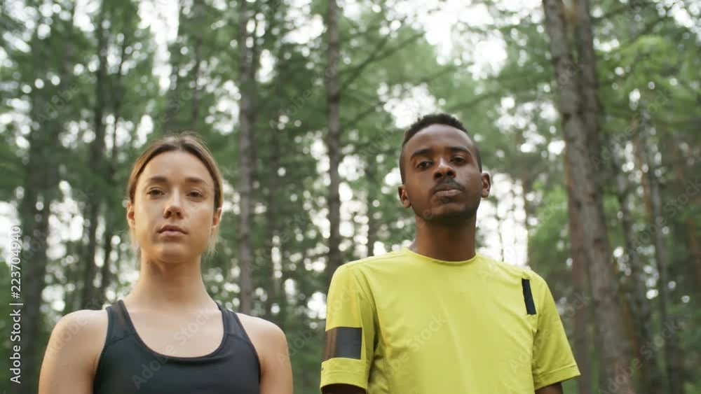 Low angle tracking of young woman in sports top and black man standing in forest and looking at camera with confidence Low angle tracking of young woman in sports top and black man standing in forest and looking at camera with confidence