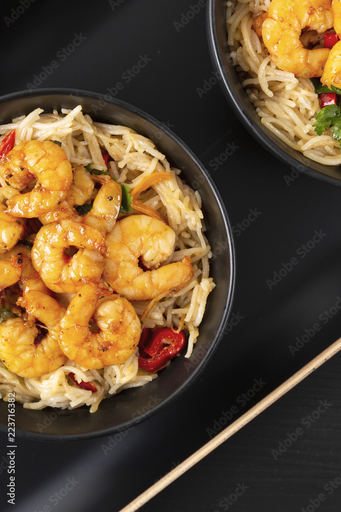 Fried prawns and rice noodles with spices on a black plate. Black background. Eastern food. Copy space. Vertical shot. Fried prawns and rice noodles with spices on a black plate. Black background. Eastern food. Copy space. Vertical shot.