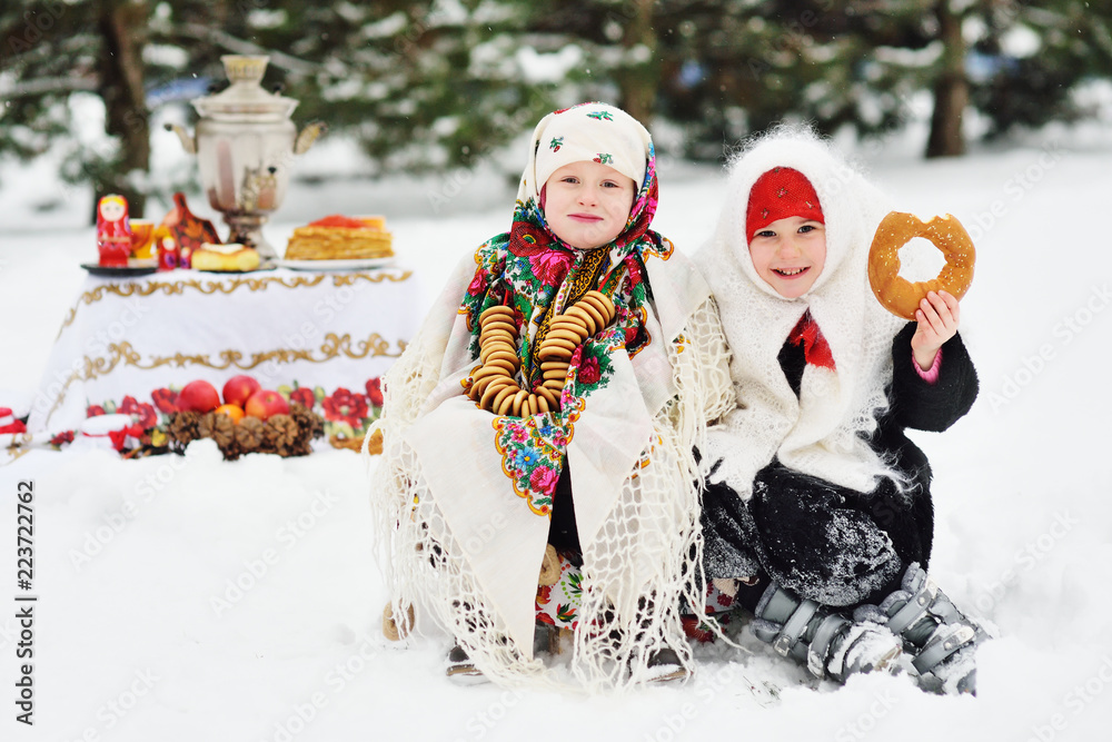 two little girls in fur coats and shawls in Russian style on the background of pancakes with red caviar, bagels, samovar 