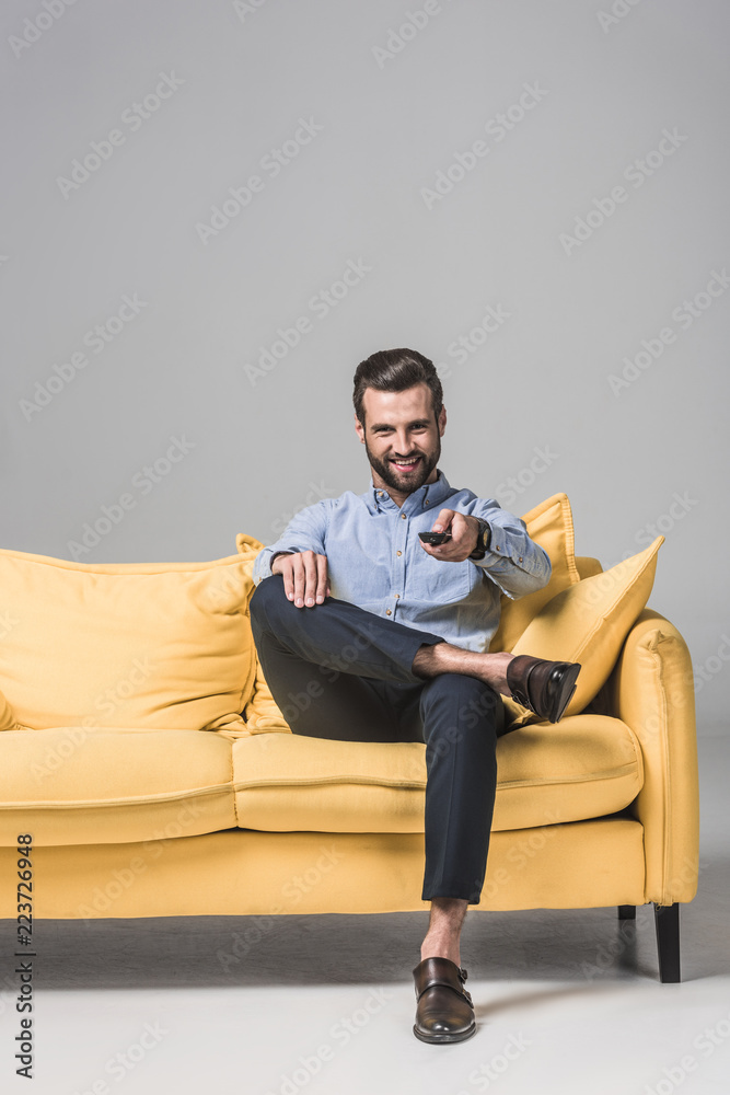 cheerful bearded man with remote control watching TV and sitting on yellow sofa on grey