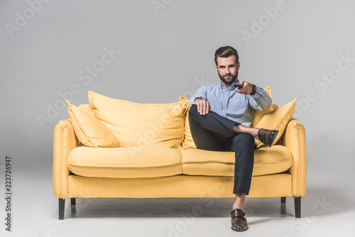handsome smiling man with remote control watching TV and sitting on yellow sofa on grey