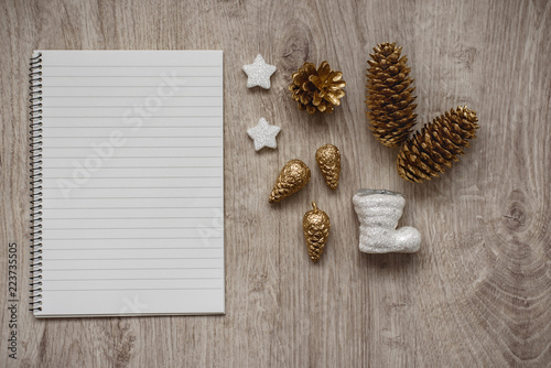 Christmas decorations, Santa's white boot, golden fir cones, snowflakes and an open blank notebook, wooden background