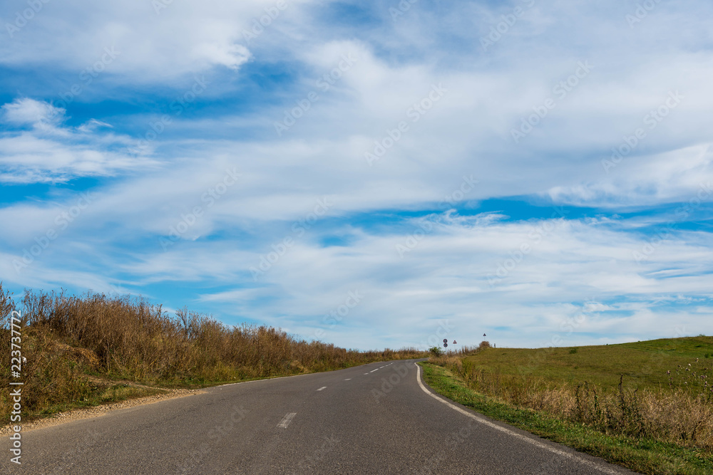 Empty asphalt road, dried vegetation, blue sky with washed white clouds.