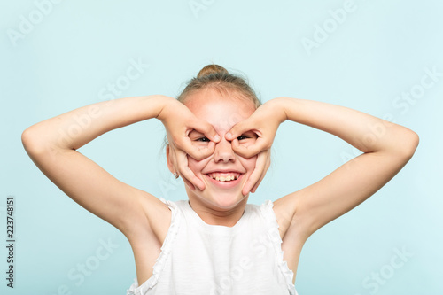 funny ludicrous joyful comic playful girl pretending to look through binoculars made of hands. portrait of a kid on blue background. emotion facial expression concept.