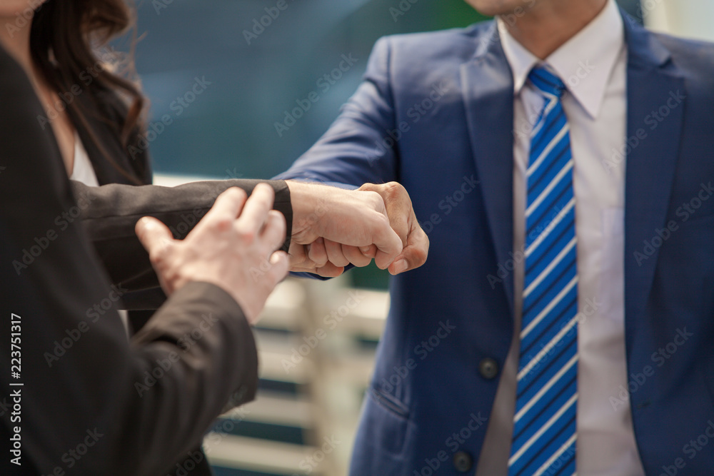 Close up of businessman making a fist bump on building with young woman in background. Business people wear suit do a fist pump together after good deal. Business success and teamwork concept.