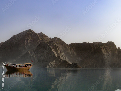 A traditional dhow boat sits in front of rocky mountains in the calm waters of Khor Ash Sham in the fjords of the Musandam Peninsula in northern Oman