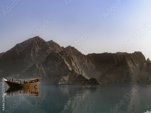 A traditional dhow boat sits in front of rocky mountains in the calm waters of Khor Ash Sham in the fjords of the Musandam Peninsula in northern Oman photo
