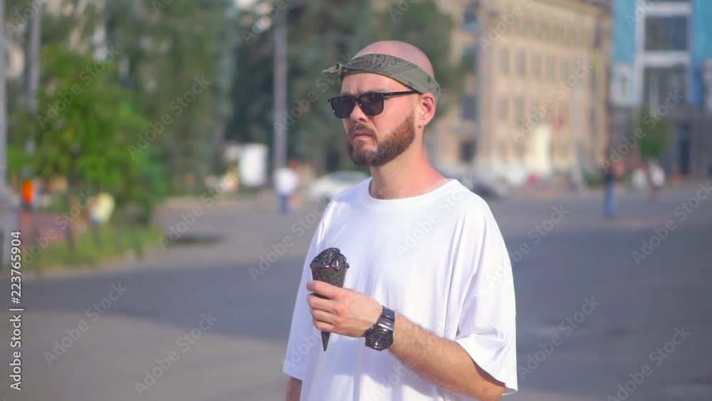 Young man in sunglasses and a white T-shirt standing on the street and holding an ice cream cone in his hands Young man in sunglasses and a white T-shirt standing on the street and holding an ice cream cone in his hands
