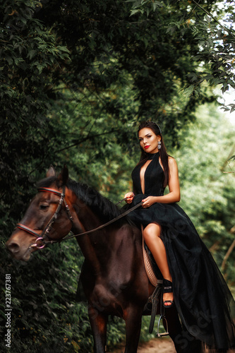 A brunette girl in a black dress on a horse in the woods.