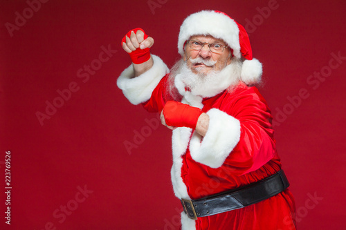 Christmas. Santa Claus with red bandages wound on his hands for boxing imitates kicks. Kickboxing, karate, boxing. Isolated on red background.