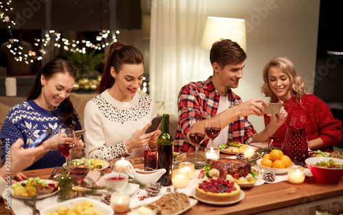 holidays and celebration concept - happy friends with smartphones having christmas dinner at home
