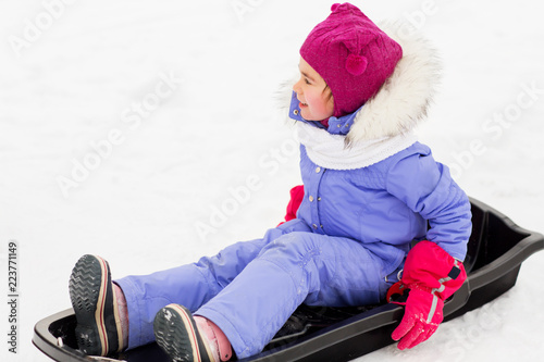 childhood, sledging and season concept - happy little girl in snowsuit sitting in sled outdoors in winter photo