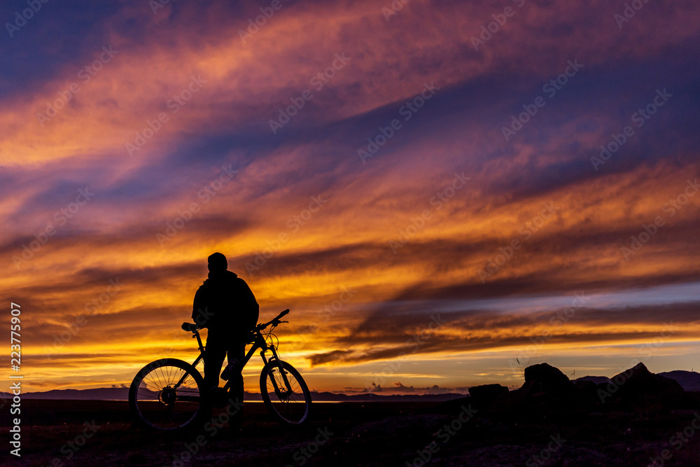 Fototapeta premium Silhouette of a cyclist on the background of a magnificent sunset. The guy is standing with the bike and watching the sunset