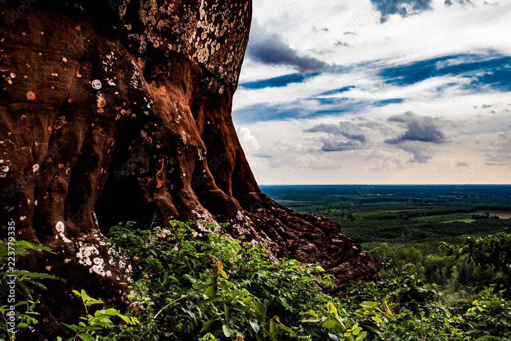 A red rock cliff side high above the ground. Rain forest and deep skies make up the background.