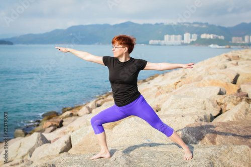 adult woman with short red hair in sportswear doing yoga exercise outdoors with sea views