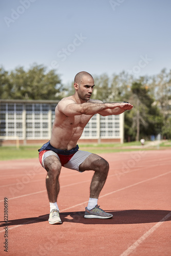one young muscular man, shirtless, side view, squat exercise. outdoors, sports venue, running tracks.