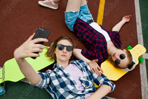 Two pretty blond girls wearing checkered shirts, caps and denim shorts are lying on the bright longboards on the sportsfield with their sunglasses on and making selfie. Sport and cool style. photo