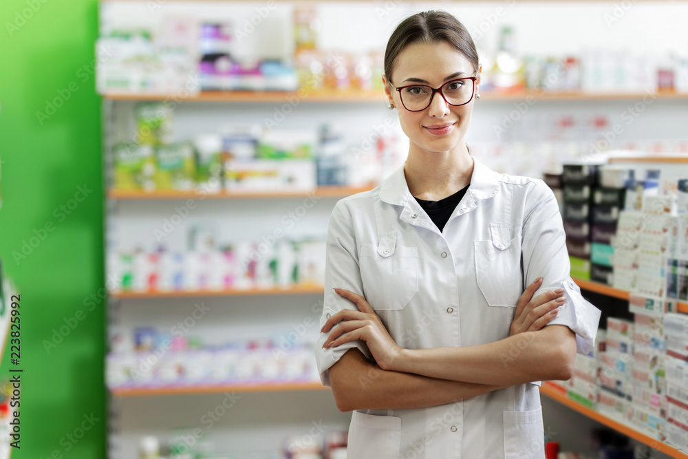 A smiling slim dark-haired lady with glasses, wearing a white coat, stands next to the shelf in the modern pharmacy. A smiling slim dark-haired lady with glasses, wearing a white coat, stands next to the shelf in the modern pharmacy.
