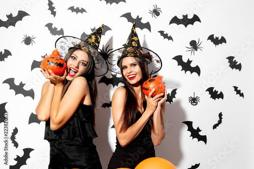 Two charming brunette women in black dresses and witches hats hold Halloween pumpkins on a white background with bats and spiders