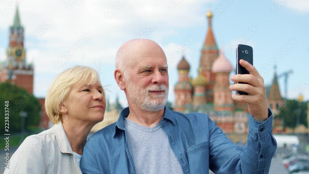Snior couple stand on Basil's Descent do selfie on mobile phone. Traveling pensioners make photo in Moscow, Russia. Red Square, St. Basil's Cathedral and Kremlin clock on background. Hand-held camera Snior couple stand on Basil's Descent do selfie on mobile phone. Traveling pensioners make photo in Moscow, Russia. Red Square, St. Basil's Cathedral and Kremlin clock on background. Hand-held camera