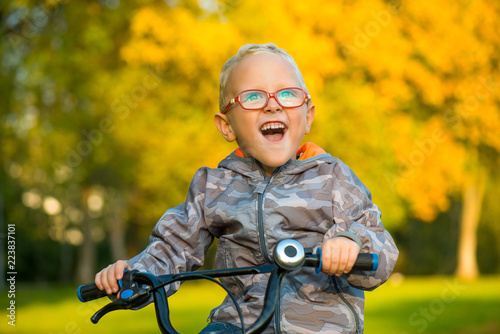Счастливый улыбающийся мальчик в очках, на велосипеде осенью в парке 