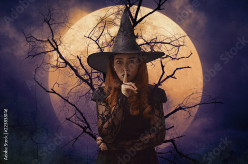 Halloween witch showing silence sign with finger over lips standing over dead tree, full moon and spooky cloudy sky, Halloween mystery concept