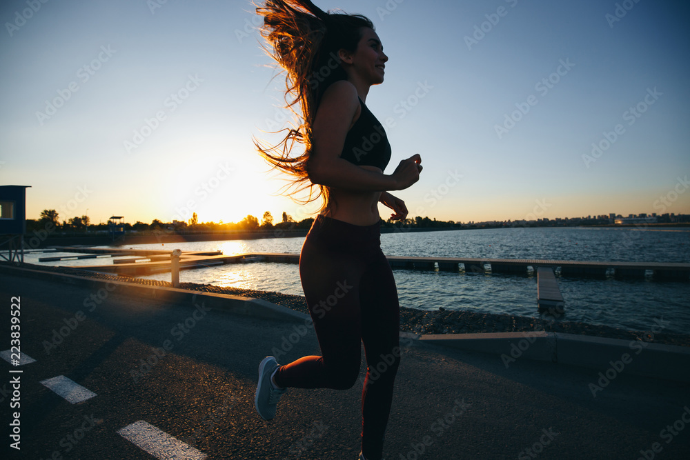 Fototapeta premium Gorgeous young girl with long brown hair dressed in sports clothes runs on the road along the reservoir on the sunset