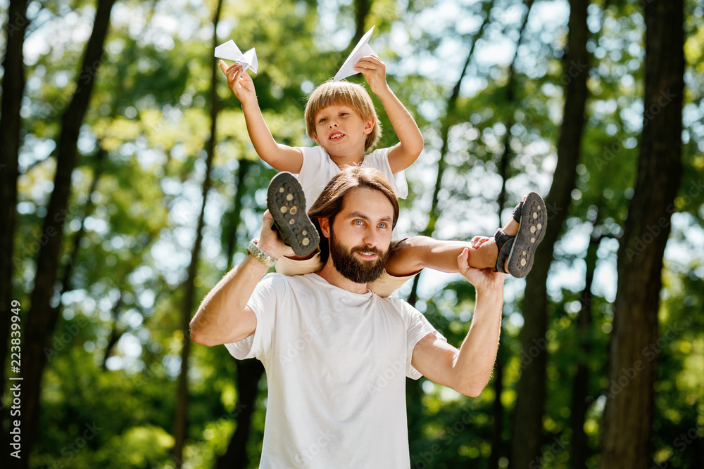 Obraz premium Little boy with his father dressed in the white t-shirts walking in the forest. Boy is sitting on the shoulders of his father and keeps paper airplanes in his hands.