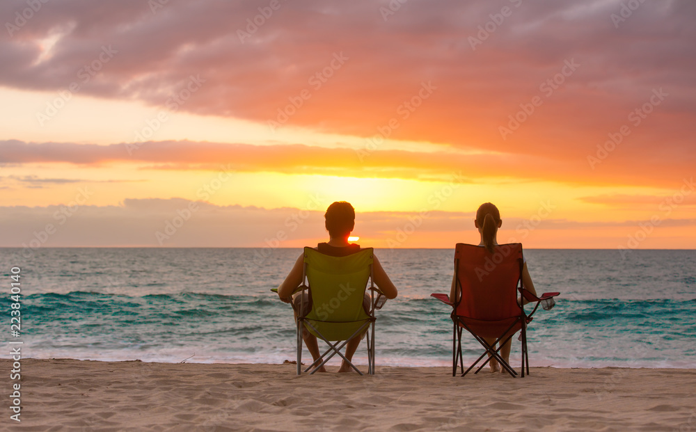 Young couple sitting on the beach relaxing watching the colorful sunset. 