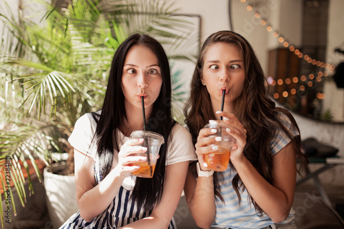 Two young smiling beautiful slim girls with long dark hair,wearing casual clothes, sit next to each other and look at the camera in a modern coffee shop.