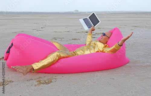 Frilanser freak. A young handsome bald man in gold metal clothes and a suit is lying on a pink inflatable sofa chair and working at the computer. The man won the lottery or the stock market photo