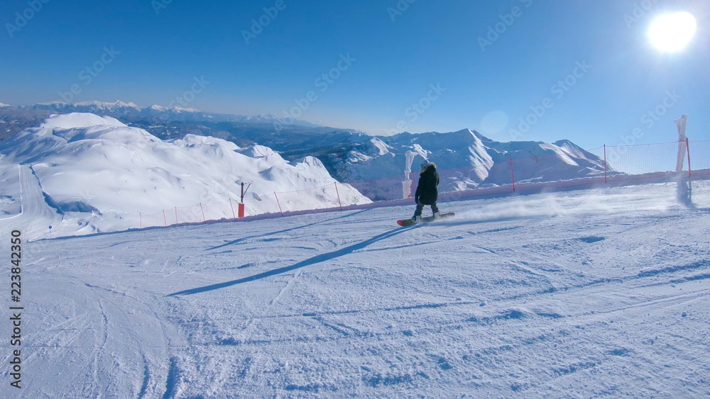 FOLLOW: Bright winter sun shines on woman snowboarding down an empty slope.