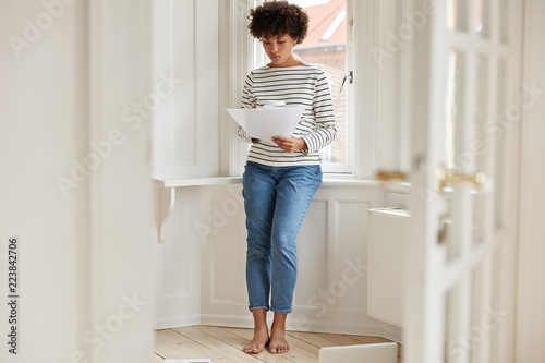 Horizontal shot of pleasant looking busy young woman studies provision for taxation, holds papers, dressed in casual clothes, stands near window sill in spacious light room, ratifies contract photo