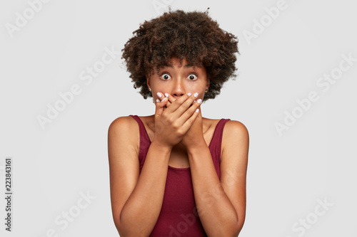 Shocked Afro American woman tries be speechless and mute, keeps hands on mouth, recieves bad news from interlocutor, wears casual outfit, stares with bugged eyes at camera, isolated over white wall